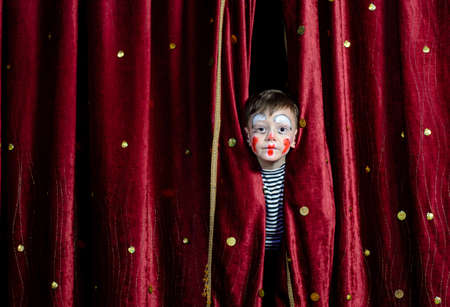 Young Boy Wearing Clown Make Up Peering Out Through Opening in Red Stage Curtainsの写真素材