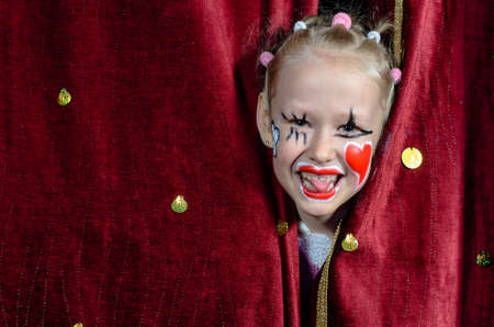 Young Blond Girl with Face Painted in Clown Make Up Smiling and Peeking Out Through Opening in Red Stage Curtainsの写真素材