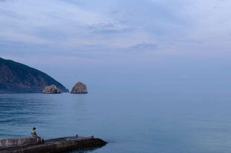 Lonely man sitting on a coastal pier looking out over a calm misty ocean with a headland visible in...