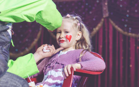 Young Blond Girl Having Face Painted by Adult While Sitting in Chair on Stage with Red Curtainの写真素材