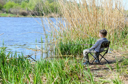 Young Boy Sitting on a Chair at the Riverside, Holding his Fishing Rod While Catching Fish at the River.の写真素材