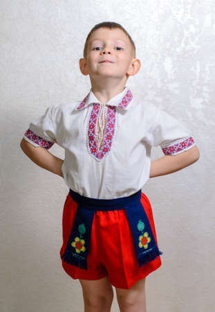 Ukrainian cute boy posing with hands behind back proud to wear the traditional folk costume with short pants and handmade embroideries on vest, white shirt and belt, portrait with copy space on greyの写真素材