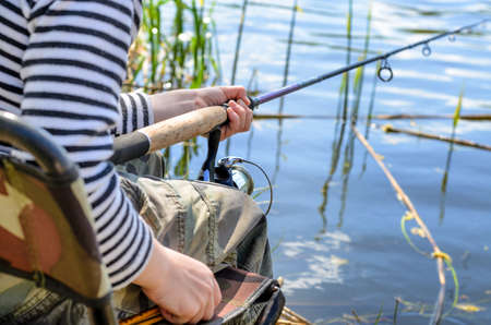 Close up view of the hands, rod and reel of a young boy sitting in a chair fishing at a tranquil...