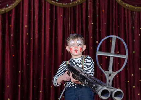 Young Boy Wearing Clown Make Up Standing on Stage Aiming Large Prop Shot Gun Rifle in front of Red Curtainの写真素材