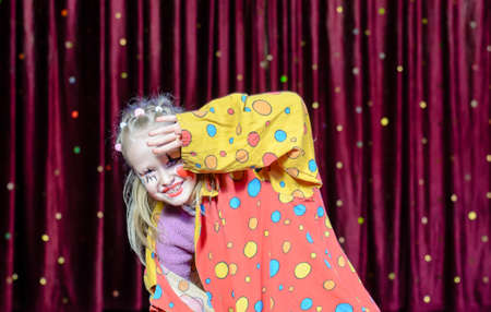 Young Blond Girl Wearing Clown Make Up and Polka Dot Costume Shielding Eyes from Bright Stage Lights in front of Red Curtainの写真素材