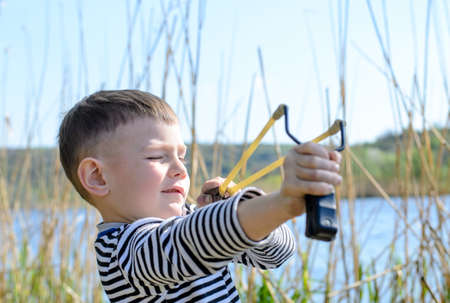 Young Smiling Boy Wearing Striped Shirt Holding Sling Shot with Rubber Bands Drawn Back in Anticipation of Catapulting Projectile Outdoors Near Lakeの写真素材