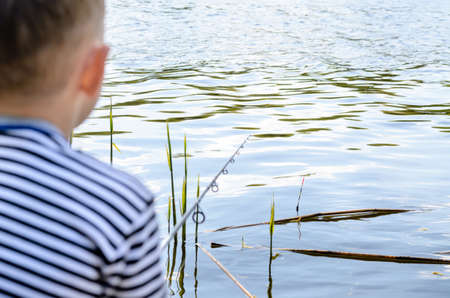 Rear View of Young Boy Sitting in Folding Chair at Edge of Pond and Fishing Patiently Amongst the Reedsの写真素材