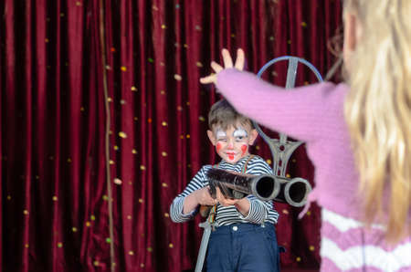 Young Boy Wearing Clown Make Up Smirking and Aiming Large Shot Gun at Blond Girl Holding Hands Up in Protest in Foreground on Stage in front of Red Curtain, Social Commentary on Youth and Violenceの写真素材
