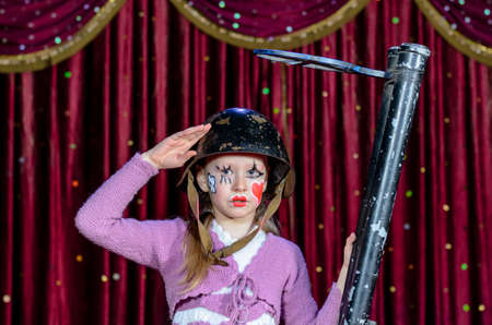 Full Length Portrait of Girl Wearing Clown Make Up, Striped Dress and Helmet Holding Over Sized Gun on Stage in front of Red Curtainの写真素材
