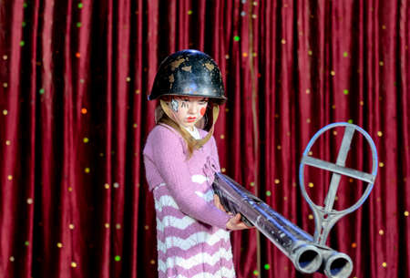 Serious Young Blond Girl Wearing Clown Make Up and Military Helmet Aiming Over Sized Rifle Gun Toward Camera and Standing on Stage in front of Red Curtainの写真素材