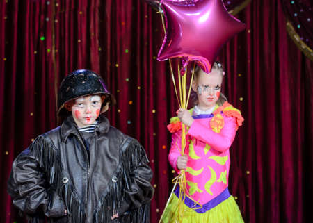 Portrait of Young Male Clown Wearing Leather Jack and Helmet Standing with Young Female Clown Wearing Brightly Colored Costume and Holding Balloons on Stage with Red Curtainの写真素材