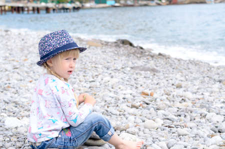 Young Blond Girl with Hair in Braids Sitting on Rocky Beach Wearing Floral Print Hat and Looking Worriedの写真素材