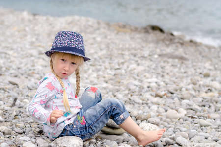 Young Blond Girl with Hair in Braids Sitting on Rocky Beach Wearing Floral Print Hat and Looking Out at View of Oceanの写真素材