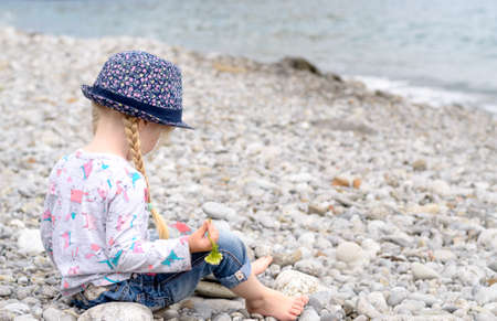 Young Blond Girl with Hair in Braids Sitting on Rocky Beach Wearing Floral Print Hat and Looking Out at View of Oceanの写真素材
