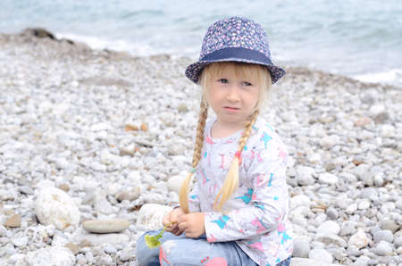 Young Blond Girl with Hair in Braids Sitting on Rocky Beach Wearing Floral Print Hat and Looking Worriedの写真素材