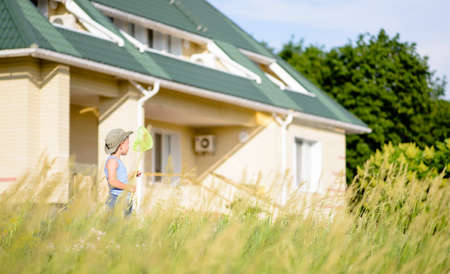 Young Boy Holding Bug Net Standing in Long Grass on Lawn of Modern House and Looking Away from Camera into the Distanceの写真素材