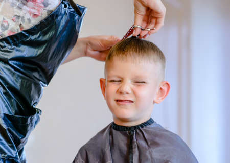 hairdresser trimming a little boys hair with a pair of scissors as her pulls a comical face with his eyes closedの写真素材