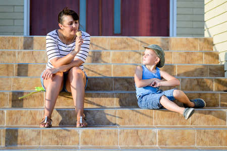 Young Boy Enviously Watching Mother Eat Ice Cream Cone While Sitting on Steps in front of Homeの写真素材