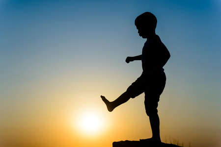 Little boy silhouetted on a wall or rock kicking the fiery orange orb of the setting sun with his foot, with copyspaceの写真素材
