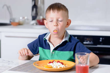 Young Blond Boy with Mouth Stuffed Full Eating Cheese and Fruit Off Orange Plate While Sitting at Kitchen Table with Glass of Red Juiceの写真素材