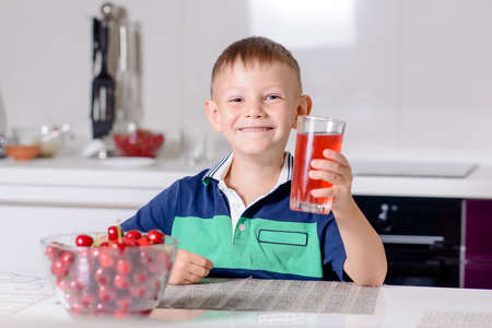 Portrait of Smiling Young Boy Drinking Glass of Red Juice While Sitting at Table with Bowl of Cherries in Kitchenの写真素材