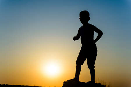 Small boy silhouetted by a colorful sunset standing on a rock or wall facing the glowing fiery sun in a twilight skyの写真素材
