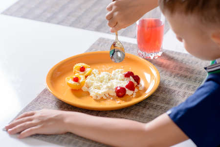 Looking Down at Young Boy Eating Plate of Cheese and Fruit with Spoon While Sitting at Dining Table with Placematsの写真素材