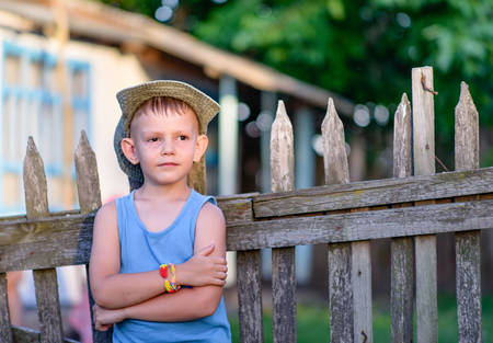 Cute Young Boy Leaning Against the Wooden Fence with Arms Crossed Over his Stomach, Showing a Pensive Facial Expression.の写真素材