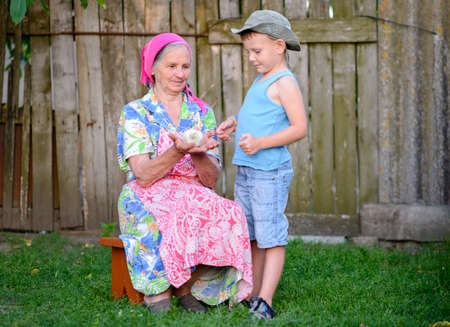 Young Boy with Grandmother in Traditional Clothing Holding Young Fuzzy Chick Together Outdoors in Yard on Rural Farm Propertyの写真素材