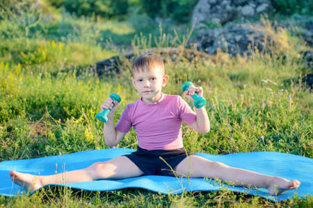 Smiling Cute Male Kid Lifting Small Dumbbells While Splitting his Legs on Fitness Mat at the Grassy Ground.の写真素材
