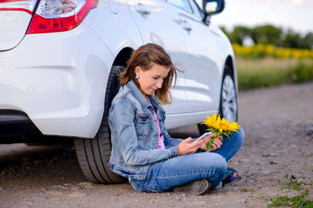 Attractive Girl with Mobile Phone Looking at the Camera While Sitting on the Ground with Sunflower Beside the White Car.の写真素材