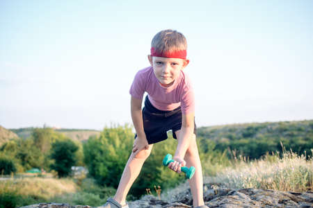 Cute Young Boy Lifting Dumbbell While Leaning his Body Forward on Top of a Boulder and Smiling at the Camera.の写真素材