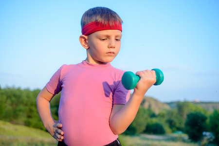 Close up Strong Cute Boy with Red Headband Doing an Outdoor Exercise and Lifting Small Dumbbell Against Blurry Nature Background.の写真素材