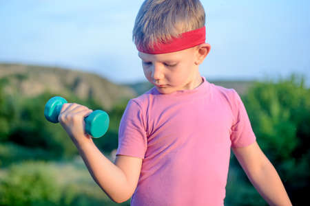 Close up Thoughtful Cute Young Boy with Red Headband Lifting Small Dumbbell While Looking Down Against Blurry Nature Background.の写真素材
