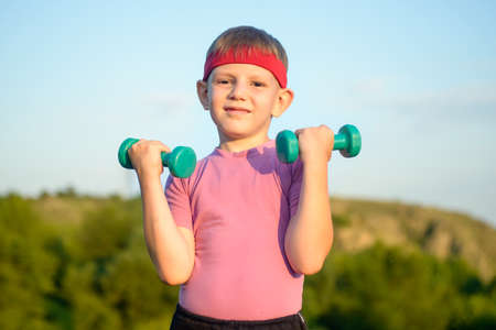Half Body Shot of an Athletic Cute Boy with Red Warrior Headband, Lifting Two Small Dumbbells and Looking at the Camera Against Green Mountain and Sky.の写真素材