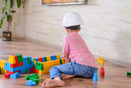 Young boy wearing hardhat playing indoors sitting on a wooden floor grinning as he holds up a large colorful plastic building blockの写真素材