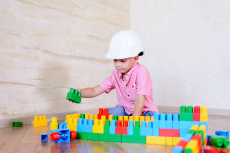 Young boy wearing hardhat playing indoors sitting on a wooden floor grinning as he holds up a large...