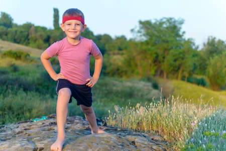 Happy little boy working out in nature standing bare foot on a rock doing stretching exercises and smiling at the cameraの写真素材