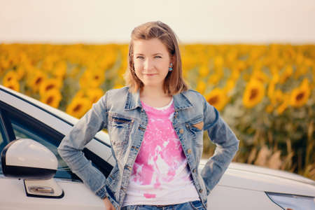 Young girl standing leaning on the bonnet of a car parked in the countryside in front of a field of...