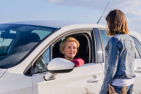 Friendly teenage girl wearing blue denim jacket and jeans while talking with a female driver stopped on a road in the countryside, in a sunny day of summerの写真素材