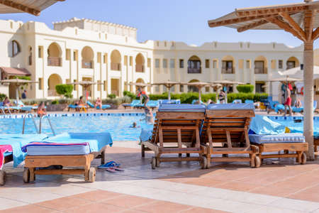 Swimming pool at a tropical resort with people swimming in the cool blue water and umbrellas and recliner chairs on the paved surround with a hotel or holiday apartment complex behindのeditorial素材