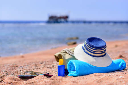 Beach Bag and Supplies for Day at the Beach on Shore with Deserted Beach in Background - Sun Hat, Flip Flops, Towel, Sunscreen Lotion, and Beach Bag on Sunny Beachの写真素材