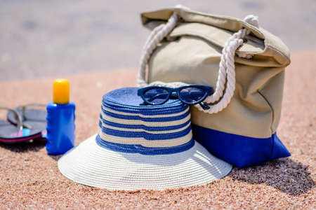 Beach gear on the sand overlooking the sea with a sunhat, sunscreen, slip slops and beach bag conceptual of a summer vacation in the tropicsの写真素材