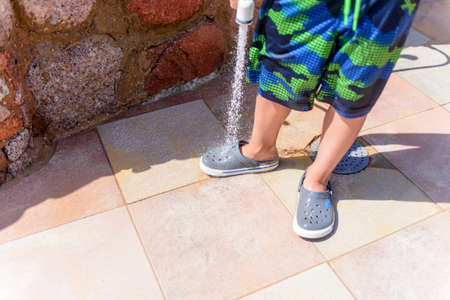 Young boy hosing the beach sand off himself and his shoe using a hosepipe at a tropical resort after...