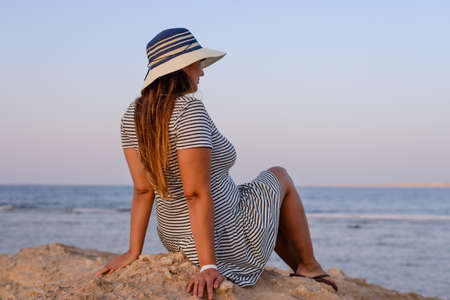 Romantic young woman wearing hat, striped dress and flip flops while sitting on the shore enjoying the breeze at the seaside in the morningの写真素材