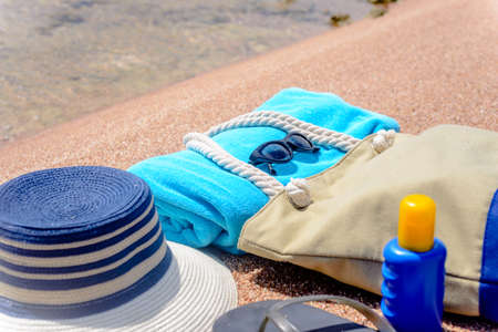 Assorted beach accessories on the sand of a tropical beach overlooking the water with a sunhat, sunscreen, towel, thongs, sunglasses and beach bagの写真素材