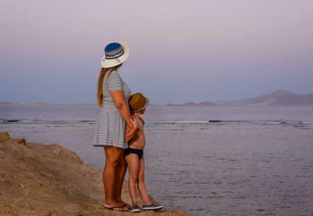 Mother with her young son on a sunset beach standing at the edge of the sea looking out over the horizon on a perfect summer dayの写真素材