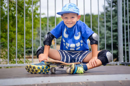 Cute happy little boy skateboarder sitting on a sidewalk with his skateboard in front of him in his protective gear grinning at the cameraの写真素材