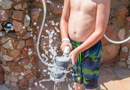 Boy spraying off his shoes with water from a hosepipe to wash out the sand from the beach at an outdoor facility at a tropical seaside resortの写真素材