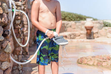Boy spraying off his shoes with water from a hosepipe to wash out the sand from the beach at an outdoor facility at a tropical seaside resortの写真素材